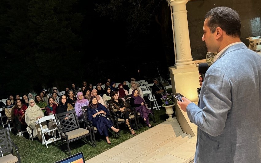 A person standing on a stage with a microphone and a group of people sitting on chairs

Description automatically generated