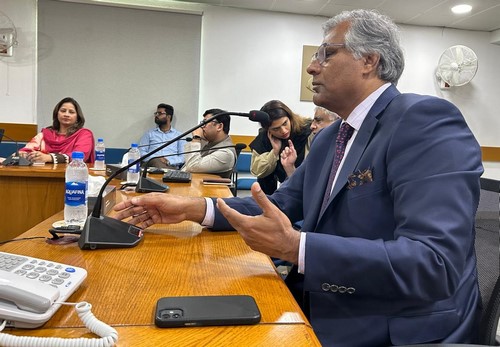 A person in a suit sitting at a table with a microphone

Description automatically generated