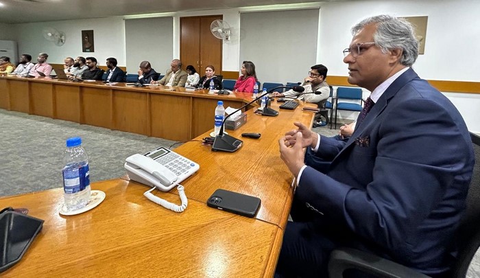 A person in a suit sitting at a table with people in the background

Description automatically generated