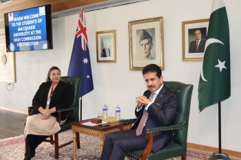 A person in a suit sitting in a chair with a person in a room with flags

Description automatically generated