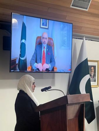 A person in a white head scarf sitting at a podium with a microphone and a screen with flags

Description automatically generated